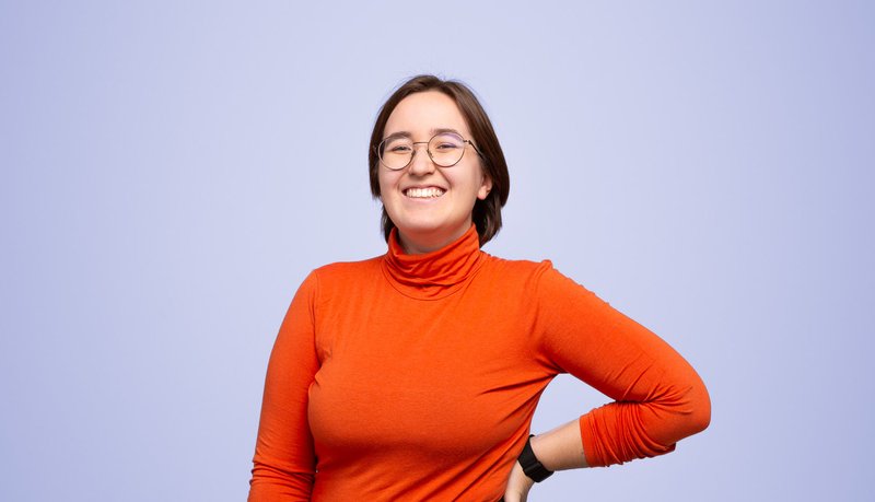 Portrait von Paula Vogel, Digital Creation bei Communicon. Sie steht vor einer lila Wand, lächelt freundlich in die Kamera und trägt einen roten Pullover.
