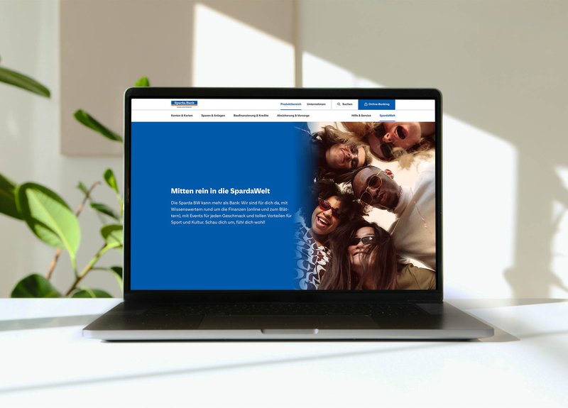Laptop auf einem Tisch zeigt die Website der Sparda-Bank mit großem Banner („Mitten rein in die SpardaWelt“) und einer Gruppe junger Menschen.